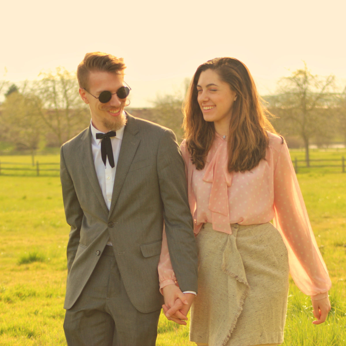 A couple holding hands and walking arm-in-arm through a grassy field at sunset. The man wears a gray suit, white shirt, black bow tie, and sunglasses.